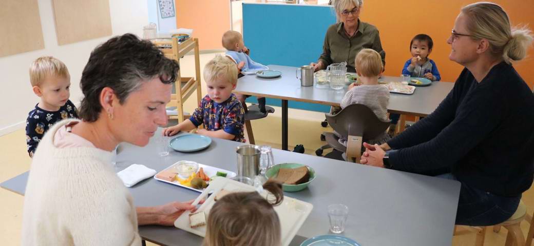 Voksen hjælper vuggestuebarn ved frokostbord