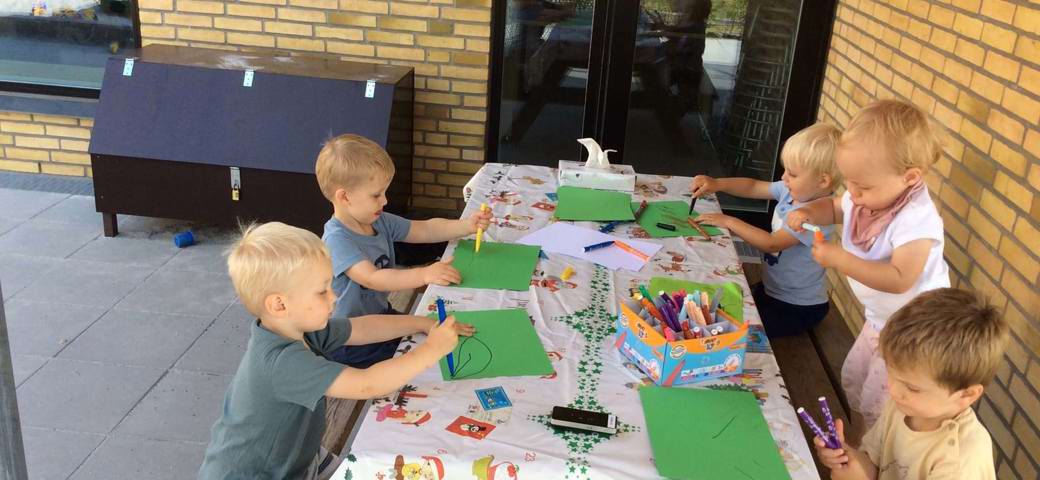 Børn tegner omkring bord i skyggen