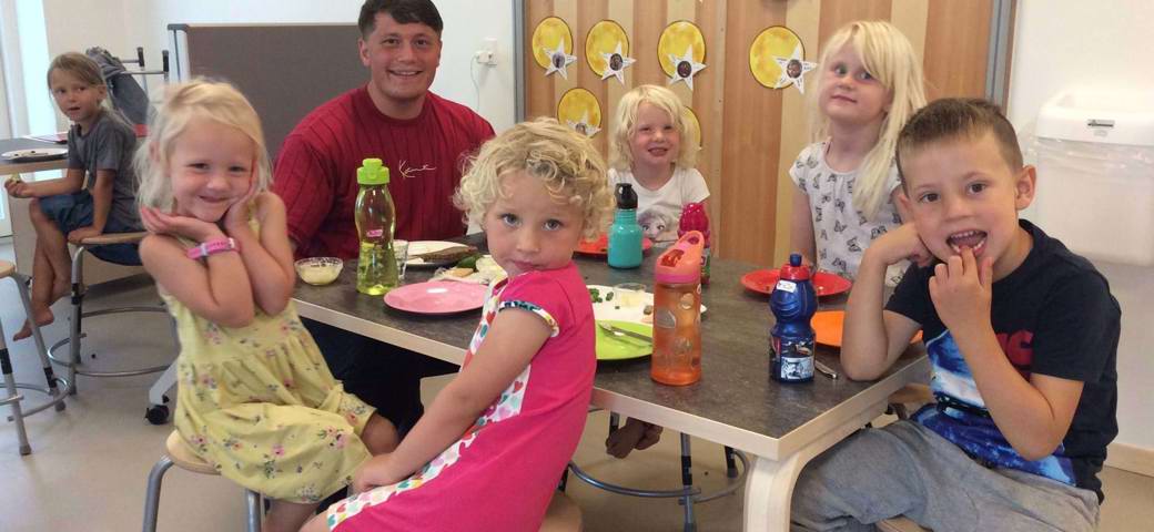 Glade børn venter på frokost omkring bord indenfor